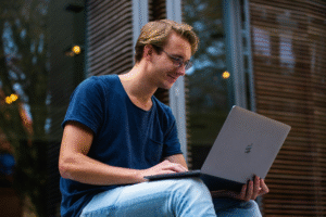 A man sitting on a bench, focused on his laptop, surrounded by greenery in a park setting.